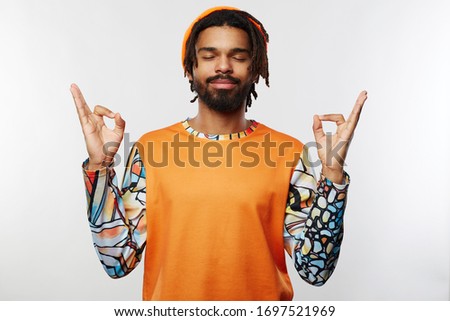 Pleasant looking young unshaved brunette guy with dark skin folding hands in mudra gesture and keeping his eyes closed while standing over white background