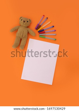 Childish coloring, painting and educational concept image of a blank mockup page, a soft brown teddy bear and some colorful crayons, top view on orange background