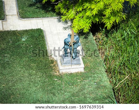 Small lizard is on the miniature statue of two soldiers surrounded by grass and bushes. Lizard is sitting on top of the soldiers which are statues.