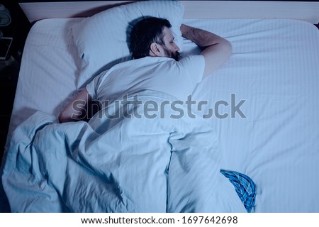 A lonely bearded dark-haired man sleeps soundly lying on his stomach at night on the bed, hands clasped. The general plan from above, white bedding, a bedroom, self-isolation. One Royalty-Free Stock Photo #1697642698