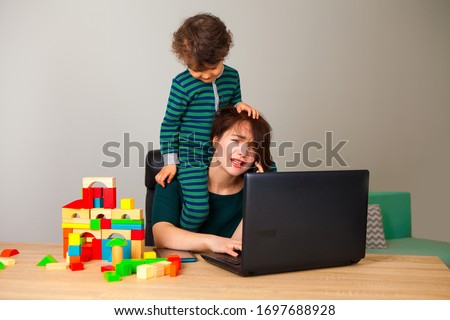 work at home. tired woman with a child on his neck sitting at a computer and talking on the phone with the employer while the child is playing cubes and hanging around her. inability to work at home. Royalty-Free Stock Photo #1697688928