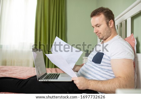 Adult man lying in his bed while he is working from home on his laptop. Papers or bills in his hand and a cheersful face