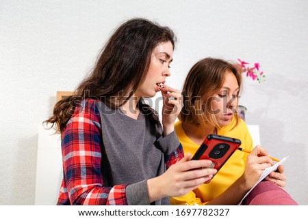 Two young Caucasian women are resting in the bedroom, using a smartphone and writing something carefully on paper. Concept of LGBT relations and social networks