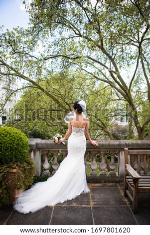 Picture of elegant bride wearing white dress with train and stylish veil standing back on the terrace and holding round wedding bouquet, nice green spring trees on the background