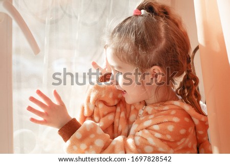 A little girl in plush pajamas with a hood plays on the window behind the curtain. A girl walks at home on a window during a coronavirus epidemic in self-isolation.