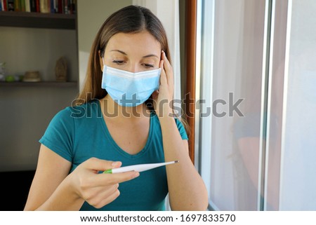 COVID-19 Pandemic Coronavirus Surgical Mask Woman Checking Temperature with Thermometer at Home Symptom of SARS-CoV-2. Girl with mask on face check fever one symptom of Coronavirus Disease 2019. Royalty-Free Stock Photo #1697833570