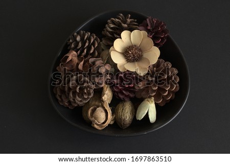 closeup of pot potpourri in a  brown rounded corner triangular ceramic dish against a black background. A wooden flower and pine cones are in the dish