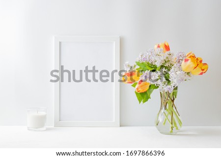 Home interior with decor elements. Mockup with a white frame yellow tulips and branches of lilac in a vase on a light background