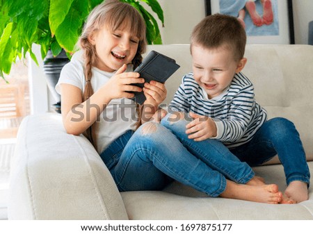 kids are watching cartoons on smartphone indoors. brother and sister are sitting on sofa in living room at home. laughing happy  kids faces.