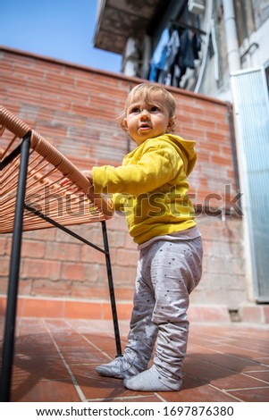 cute baby girl with blond hair and yelllow hoodie playing in a garden patio with a chair