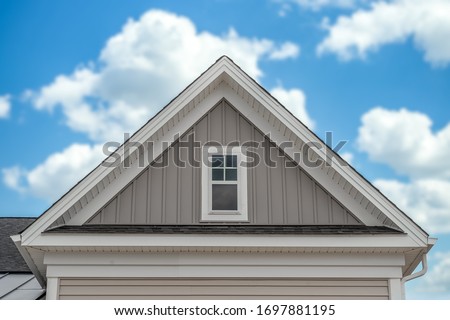 In focus gable on a newly built single family house with white fascia, common rafter, soffit, vertical vinyl siding, decorative white trim, rectangular double sash attic window blurred cloudy sky Royalty-Free Stock Photo #1697881195