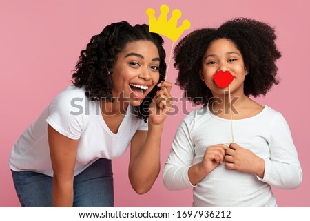 Portrait of joyful black mom and little daughter holding paper party accessories, having fun over pink background, closeup