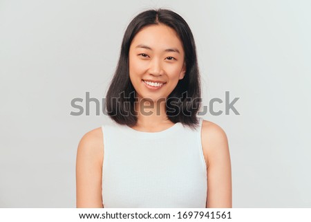 Portrait of girl tilts head and smiles sweetly showing white teeth. Beautiful young woman Asian appearance with black hair brown eyes stands isolated white background in Studio
