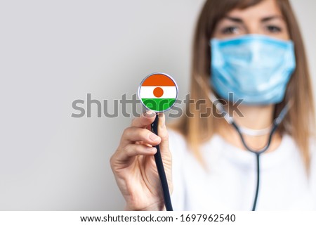 female doctor in a medical mask holds a stethoscope on a light background. Flag of Niger added on the stethoscope. Concept of medicine, level of medicine, virus, epidemic