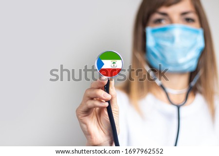 female doctor in a medical mask holds a stethoscope on a light background. Flag of Equatorial Guinea added on the stethoscope. Concept of medicine, level of medicine, virus, epidemic.