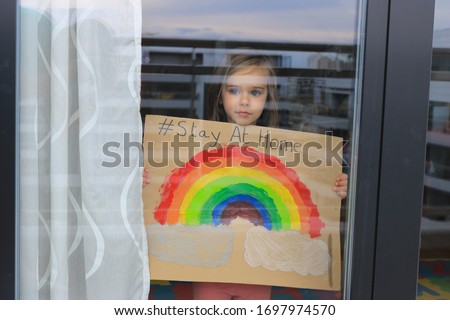 Young girl looking at infinity through a window and showing a picture of a rainbow on a cardboard, during coronavirus quarantine. Inspirational message “Stay at home”.