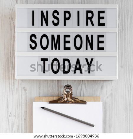 'Inspire someone today' words, clipboard with blank sheet of paper on a lightbox on a white wooden background, top view. Overhead, from above, flat lay. 