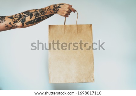 cropped photo on a hand with tattoos that holds blank craft paper package, bag mock-up. Empty space for design. White wall background 