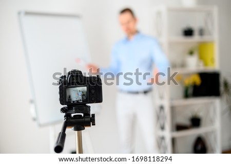 Business video blogging, vlogs about finance, sales. Young attractive guy in smart casual wear stands near a blanc flip chart board, explains something, records himself to the camera. Focus on camera