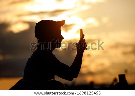 A young girl in a cap uses a smartphone to take pictures. Black silhouette against the sunset sky