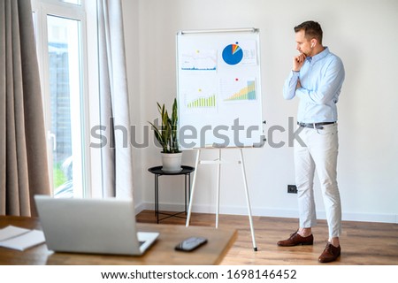 Concentrated bearded guy in a formal shirt looks at a graphs and diagrams on whiteboard, he solves business tasks, laptop in front of him. Concept of webinars, online meeting