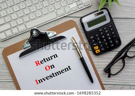 RETURN ON INVESTMENT text with notepad, keyboard, decorative vase, fountain pen, spectacles and calculator on wooden background. Business and Copy space concept