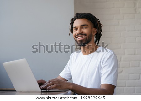 African American freelancer working from home. Portrait of young successful programmer using laptop computer, looking at camera and smiling, sitting at workplace