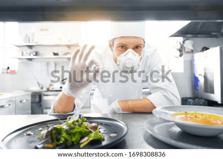 health, safety and pandemic concept - male chef cook wearing face protective mask or respirator for protection from virus disease with plate of soup and salad showing ok sign at restaurant kitchen Royalty-Free Stock Photo #1698308836
