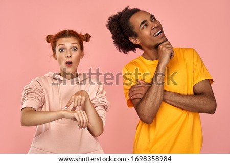 Glad dark-skinned guy focused up with smile on face, dressed in stylish clothes, shocked ginger woman indicates at wrist, demonstrates time, being in hurry, astonished to have no time for preparation Royalty-Free Stock Photo #1698358984