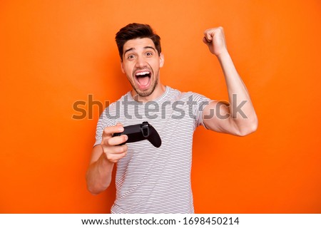 Photo of funny crazy astonished guy hold hands joystick playing video games excited gamer came finish first raise fist wear striped t-shirt isolated bright orange color background