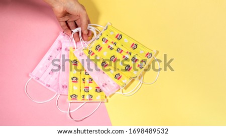 Female hand holding set of color childrens medical masks on yellow with pink backdrop. Masks - means of personal protection against virus. Pandemic Covid-2019. Top view, copy space.