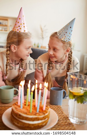 Two sisters in party hats sitting at the table with birthday cake and smiling to each other they sitting at home
