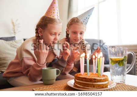 Two cute girls in party hats eating birthday cake at the table in the living room at home