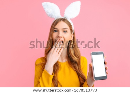 happy Easter. A shocked young woman in the ears of an Easter Bunny, holding a basket of colorful Easter eggs, shows a blank smartphone screen on an isolated pink background