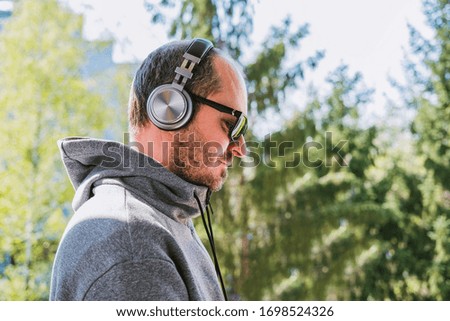 Bearded man in hoodie with headphones listening to music in park outdoors, head and shoulders