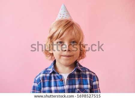 Portrait of little boy with blond hair and in party hat looking at camera over pink background