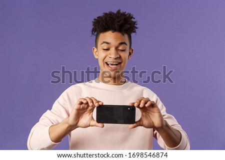 Portrait of amazed, excited young man seeing something interesting, stream concert to his internet social network profile, taking photo or recording video with mobile phone, purple background