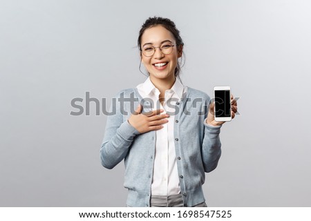 Technology, online and mobile lifestyle concept. Happy boastful asian woman showing pictures or her profile on app smartphone display, holding phone laughing and smiling camera pleased