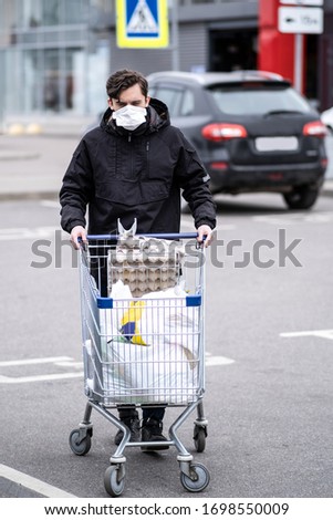 A young man in a protective mask wheeled out a cart full of groceries from a supermarket. He walks across the Parking lot to his car, about to take the groceries home.