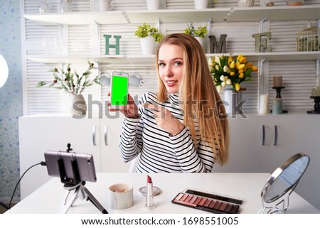 Beauty blogger woman filming advertising, holding green screen smartphone application. Make-up influencer blonde girl live streaming cosmetics product review in studio asking for likes, subscription