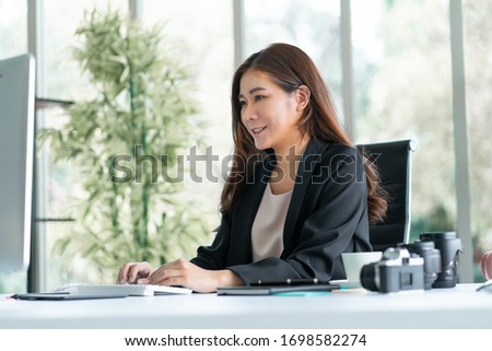 Asian businesswoman sitting at her desk in office looking at computer monitor with smile while camera and lens on her writing table. Woman entrepreneur works as digital editor photography business 