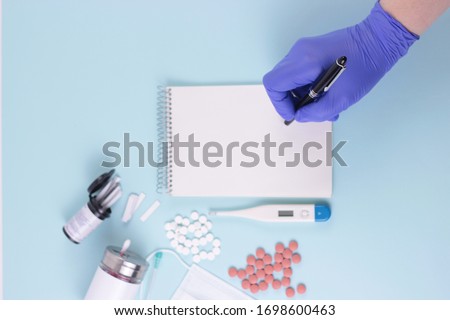 Hospital treatment, coronavirus concept.Doctor's hand in blue medical glove write a prescription.Blue desk with pills, thermometr, medicine mask, syringe.Mock up blank empty notepad.Top view overhead. Royalty-Free Stock Photo #1698600463
