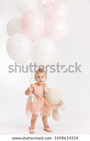 Little cute girl holds branch of balloons and large Teddy bear enjoy festive birthday event wear pink pastel summer dress isolated on a white background.