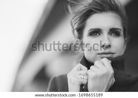 A beautiful lonely woman in a coat and scarf under a frozen bridge holds her collar with her hands. The concept of loneliness. Black and white art photo. Soft focus.