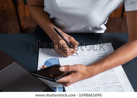 Busy young woman signing papers at comfortable office table and double checking information on mobile phone, modern business lady workplace on smart devices