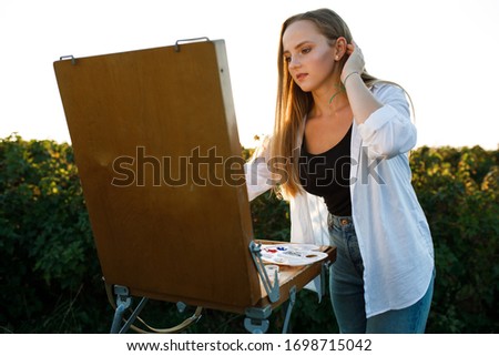 blonde girl dressed and black shirt and white shirt. girl paints a picture in nature. the girl uses colored paints and wooden brushes. girl draws a picture on a wooden easel and smiles. 