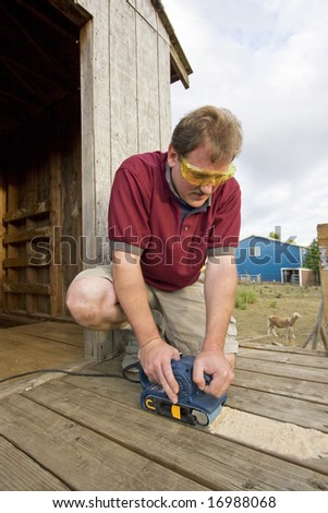 Smiling man with safety glasses using a power sander on some wood. Vertically framed photo.