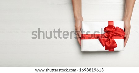 Woman holding gift box with red bow and space for text on white wooden background, top view