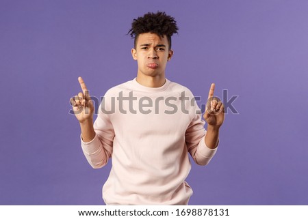 Portrait of gloomy, pouting frowning hipster guy dont have something he wants, pointing fingers up at super cool expensive thing, asking for it, trying receive compassion, purple background Royalty-Free Stock Photo #1698878131