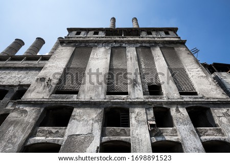Symmetrical wide angle view from below of the facade of an old stark abandoned concrete factory with four chimneys against a solid blue sky